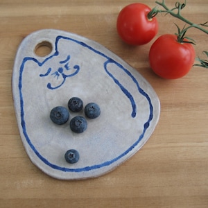 Puede incluir: Una fuente de cerámica con diseño de gato azul, con arándanos. Dos tomates rojos y una ramita de hojas verdes están sobre una superficie de madera. La fuente tiene un agujero para colgar.