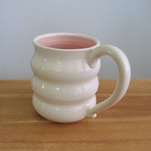 May include: A ceramic mug with a glossy off-white exterior and a soft pink interior. The mug features a distinct three-tiered, rounded, bulbous shape and a curved handle attached to the side, creating a smooth, reflective surface finish.