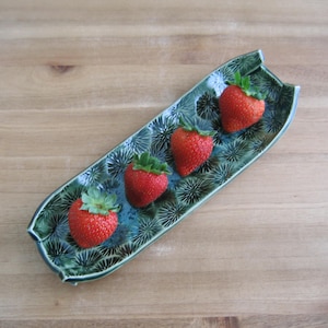 May include: A rectangular ceramic tray with curved edges and a glossy green glaze featuring a dark, radiating starburst pattern. Four fresh red strawberries with green leafy stems are arranged in a line along the center of the tray.
