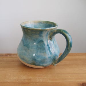 Puede incluir: Una taza de cerámica hecha a mano con un cuerpo redondeado y un asa grande. La taza presenta un esmalte azul y turquesa con un acabado brillante. El interior y la base son de color blanco crema. La taza está sobre una superficie de madera.