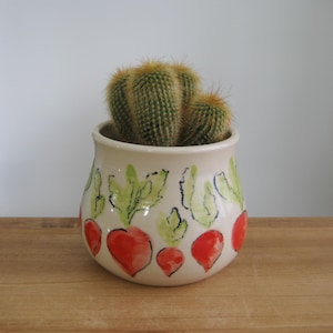 Puede incluir: Una pequeña maceta redonda de cerámica blanquecina con un diseño de rábano pintado a mano. La maceta contiene un grupo de cactus verdes. Los rábanos son rojos con hojas verdes, y la maceta está sobre una superficie de madera.