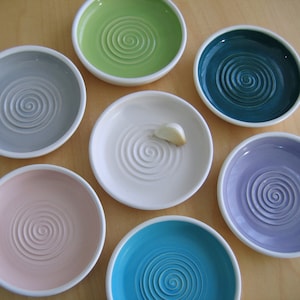 Puede incluir: Conjunto de siete platos pequeños de cerámica con borde blanco y diseño en espiral en varios colores, incluyendo azul claro, verde azulado, lavanda, rosa, gris y verde. Un plato tiene un diente de ajo.