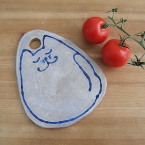 Peut inclure: Un plat de service en céramique gris clair fait à la main avec un motif de chat bleu. Le plat a une forme arrondie avec un trou en haut et un contour bleu. Deux tomates rouges sur une vigne sont à côté du plat.