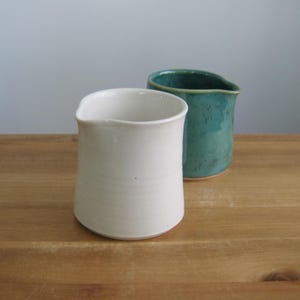 Op de afbeelding: Twee keramische kannen. Eén is wit, met een afgeronde body en een licht uitlopende rand. De andere is turquoise, met een vergelijkbare vorm en een kleine tuit. Beide staan op een houten oppervlak.