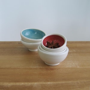 May include: Two small ceramic bowls with white exteriors and colored interiors. One bowl has a light blue interior, while the other features a red interior containing a single star anise spice. Both bowls have a smooth, rounded shape and base.