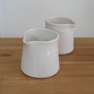 May include: Two white ceramic jugs with a pouring spout. The jugs are different sizes and sit on a wooden surface. The smaller jug is in the foreground, and the larger jug is behind it. The background is a plain white wall.