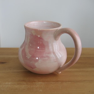 May include: A ceramic mug with a rounded, bulbous body and a curved handle. The surface features a glossy glaze with a marbled pattern of soft pastel pink and creamy white tones, creating a smooth, reflective finish across the entire exterior.