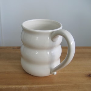 May include: A handmade, off-white ceramic mug with a unique, stacked design. The mug features a rounded handle and a bulbous body, giving it a distinctive, sculptural appearance. The mug is sitting on a wooden surface.