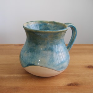 May include: A handmade ceramic mug with a rounded body and a curved handle. The mug features a blue and turquoise glaze with a cream-colored base. The mug is approximately 4 inches tall and is suitable for hot or cold beverages.