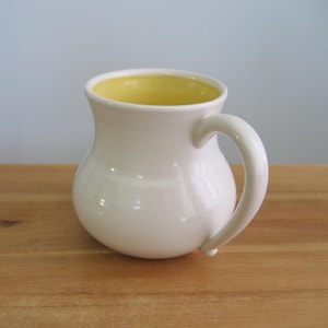 Puede incluir: Una taza de cerámica con cuerpo redondeado y abombado, con un acabado exterior blanco liso y brillante. El interior de la taza es de un color amarillo sólido y brillante. Presenta un asa curva en el lado derecho y un borde circular ancho.