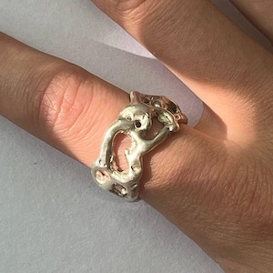 Può includere: Un anello color argento con un design organico e astratto. L'anello presenta una serie di forme irregolari interconnesse con spazi aperti, creando un aspetto unico e strutturato. L'anello è indossato su un dito.