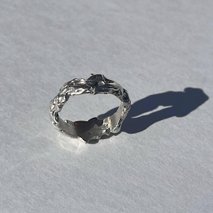 May include: A silver ring with a textured, organic design. The ring has an irregular surface, resembling a natural, sculpted form. The ring is shown on a white surface, with a shadow cast to the right.