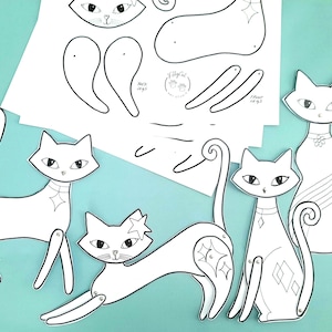 Op de afbeelding: Een verzameling witte papieren kattenuitsparingen met zwarte details, waaronder verschillende geassembleerde katten en niet-geassembleerde onderdelen. De katten hebben verschillende poses en decoratieve elementen. De achtergrond is lichtblauwgroen.