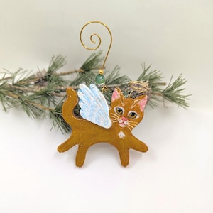 May include: A hand-painted orange cat ornament featuring light blue wings and a small gold wire halo above its head. The cat has pink inner ears, a pink nose, and white whiskers. A gold wire hook with a small green bead is attached to the top.