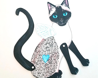 Puede incluir: Un recorte de papel con forma de gato siamés con articulaciones. El gato tiene ojos azules, marcas en blanco y negro y una cola larga y negra curvada. El cuerpo del gato presenta un corazón y alas de ángel, con el texto "LIVE" y "LAUGH".