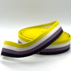 Könnte beinhalten: Gerollte Schleife in den Farben der nicht-binären Pride-Flagge. Das Band hat eine gelbe Mitte mit weißen, lila und schwarzen Streifen. Das Band besteht aus gewebtem Stoff.