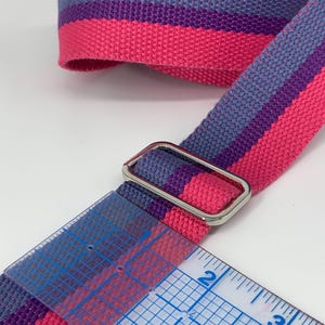 May include: A close-up of a pink, purple, and gray striped webbing strap with a silver metal buckle. The strap is measured with a ruler, showing it is 2 inches wide.
