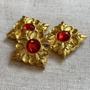Op de afbeelding: Drie gouden metalen knopen met rode edelsteen accenten. Elke knop heeft een decoratief bloemmotief.