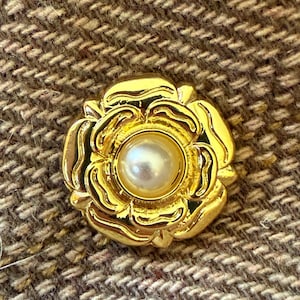 May include: A circular gold-tone metal button featuring a sculpted floral design with layered petals radiating from a central, smooth, round white pearl. The button has a polished, reflective finish and is attached to a brown and tan tweed fabric.