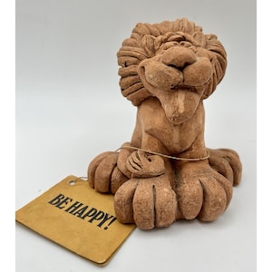 Könnte beinhalten: Terrakotta-Löwenfigur mit detaillierter Mähne, Pfoten und einem lächelnden Gesichtsausdruck. Die Figur hat ein Etikett mit der Aufschrift "BE HAPPY!" in fetten Buchstaben. Der Löwe hat eine warme, erdfarbene braune Farbe.