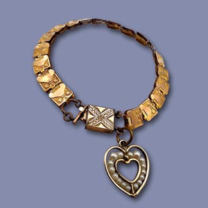 May include: A gold-toned necklace with a heart-shaped pendant. The necklace features a series of rectangular links and a square clasp. The heart pendant is outlined with small pearls, creating a delicate and elegant design.