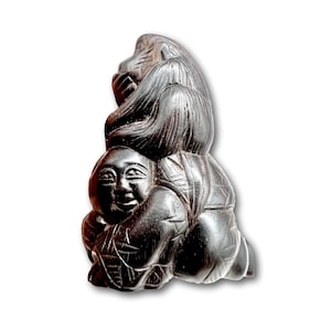 May include: A black wooden sculpture of a monkey sitting on top of a person. The monkey has a long tail and is holding its hands together. The person is sitting with their legs crossed and has a smiling face.