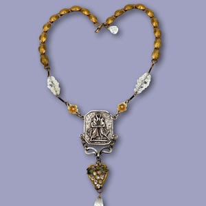 May include: An antique necklace with a heart-shaped design. The necklace features a gold-toned chain in a heart shape, with a silver pendant depicting two figures. The pendant is adorned with clear crystals and a teardrop-shaped crystal at the bottom.