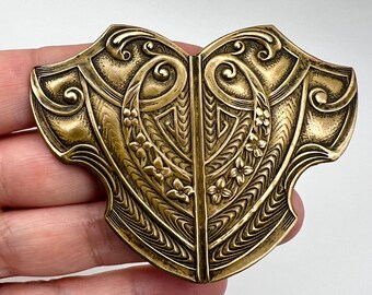 Puede incluir: Broche de metal dorado antiguo y ornamentado. El broche presenta un diseño simétrico con patrones florales y geométricos. Los bordes son curvos y detallados, con una división central. La forma general recuerda a un escudo o un corazón.