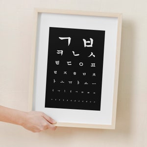 Puede incluir: Un cuadro de prueba de visión en blanco y negro enmarcado con caracteres coreanos. El cuadro está en un marco de madera claro y está siendo sostenido por una mano.