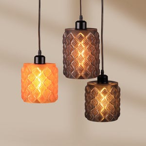 May include: Three pendant lights with textured glass shades in orange and brown tones. Each light has a cylindrical shape with a repeating diamond pattern. The lights hang from black cords and are illuminated from within, creating a warm glow.