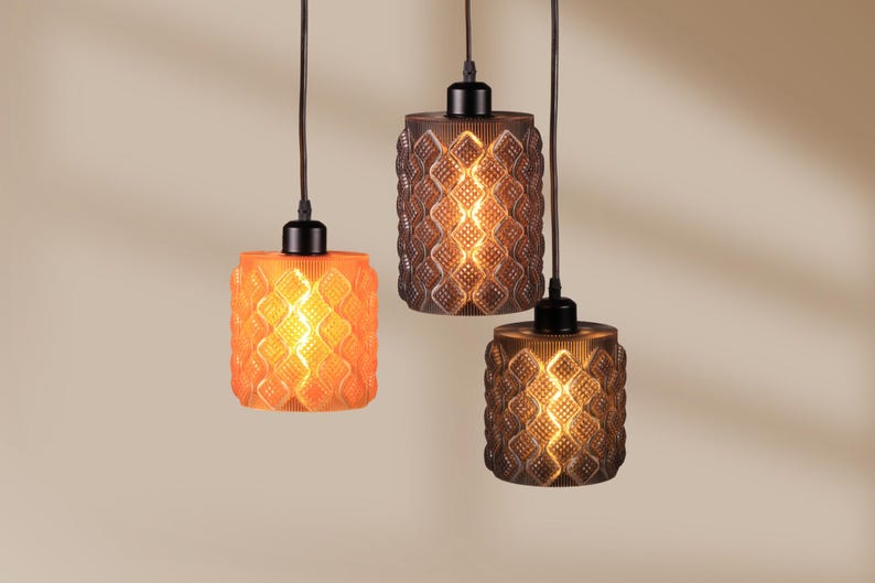 May include: Three pendant lights with textured glass shades in orange and brown tones. Each light has a cylindrical shape with a repeating diamond pattern. The lights hang from black cords and are illuminated from within, creating a warm glow.