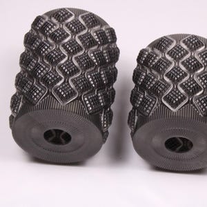 May include: Two dark gray, cylindrical objects with a textured, diamond-patterned surface. The ends have a circular design with a central hole. The objects are set against a white background.