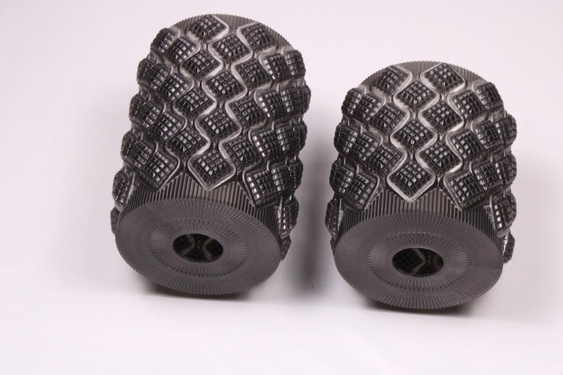 May include: Two dark gray, cylindrical objects with a textured, diamond-patterned surface. The ends have a circular design with a central hole. The objects are set against a white background.