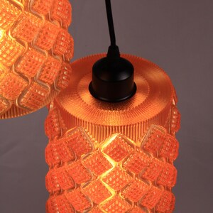 May include: Two orange pendant lights with a textured, geometric pattern. The lampshades are cylindrical with a diamond-like design. The lights hang from black cords and have black fixtures.