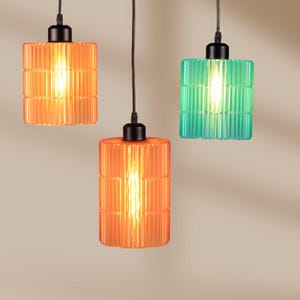 May include: Three pendant lights with cylindrical, ribbed shades in orange and teal. Each light has a black fixture and a warm, glowing bulb. The lights hang against a neutral background, creating a modern aesthetic.