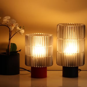 Peut inclure: Deux lampes de table en verre avec un design côtelé. Les lampes sont allumées et ont des ampoules à lumière blanche chaude. La lampe de gauche a une base rouge et la lampe de droite a une base noire.