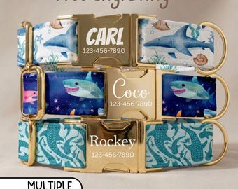 May include: Three pet collars with gold-tone metal buckles featuring custom engravings of names and phone numbers. The collars display shark patterns in white, dark blue, and teal, with each buckle showing text like Carl, Coco, or Rockey.
