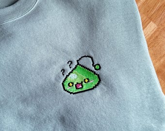 May include: A pale blue crew neck sweatshirt featuring an embroidered pixelated green slime character. The slime has a confused expression with question marks above its head. The sweatshirt is folded on a wooden surface.