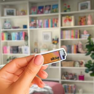 May include: A rectangular sticker with a lighter design. The sticker has a gradient of orange, yellow, and pink colors. The word "okay" is written in black font. The sticker is held in a hand.