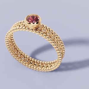 Peut inclure: Une bague en or avec une pierre précieuse rouge ronde sertie sur une bande détaillée. La bande présente un motif texturé avec de petites perles rondes et des accents délicats. La bague est présentée sur une surface violet clair.