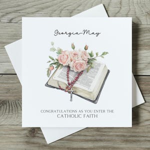 May include: A white card with the text "Georgia-May" and "Congratulations as you enter the Catholic Faith". The card features a watercolour illustration of a Bible with a rosary and pink roses.
