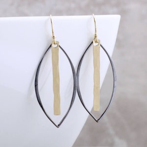 May include: A pair of gold-tone and silver-tone earrings. Each earring features a gold vertical bar suspended within a silver-tone, leaf-shaped frame. The earrings have gold-colored ear wires.
