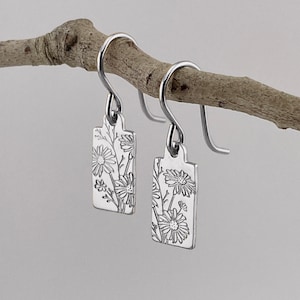 May include: A pair of silver dangle earrings with a floral design. The earrings are rectangular in shape and feature a delicate floral pattern.