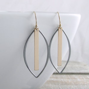 May include: A pair of gold and silver-toned earrings. Each earring features a gold vertical bar suspended within a silver-colored, leaf-shaped frame. The earrings hang from gold-colored ear wires. The background is a white surface and a wooden surface.
