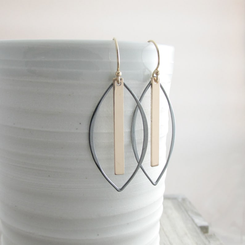 May include: A pair of gold and black earrings. Each earring features a gold vertical bar suspended within a black, leaf-shaped frame. The earrings hang from gold-colored ear wires. The earrings are displayed against a white background.