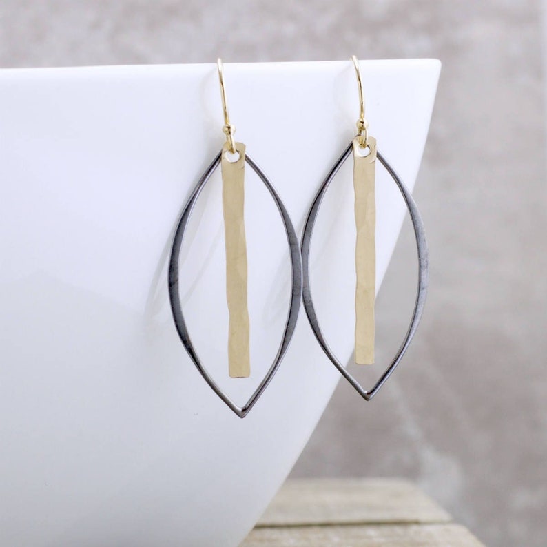 May include: A pair of gold-tone and silver-tone earrings. Each earring features a gold vertical bar suspended within a silver-tone, leaf-shaped frame. The earrings have gold-colored ear wires.