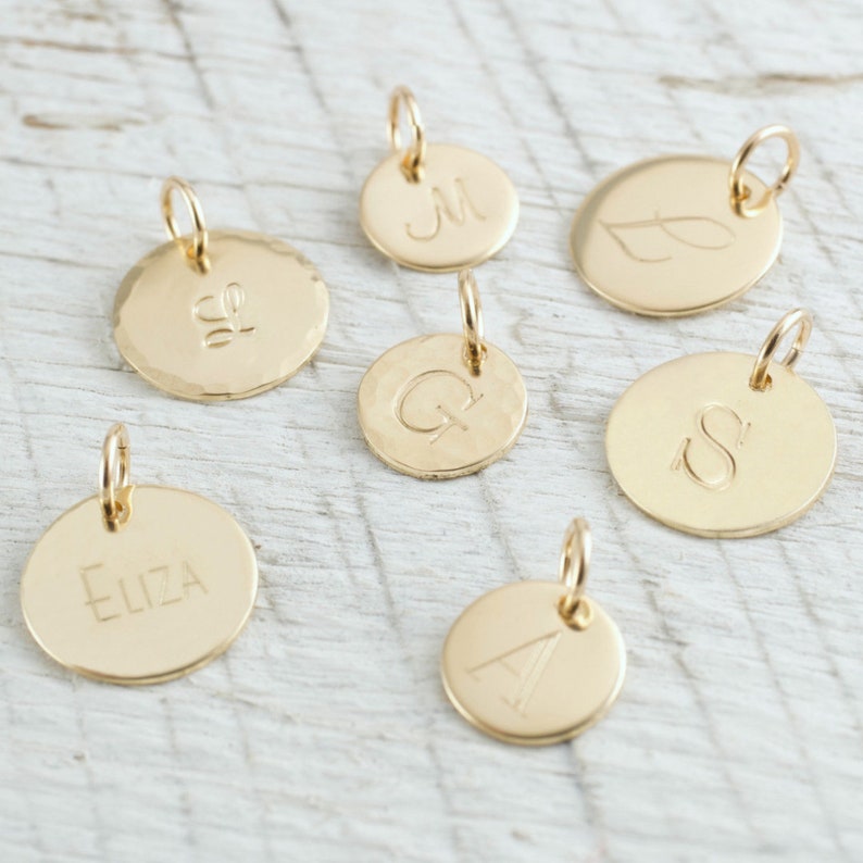 May include: Seven gold-toned metal charms with different engravings. The charms are round and have a hammered texture. The engravings include the letters "M", "S", "Q", "A", "ELIZA", and a leaf design. The charms are on a white wooden surface.