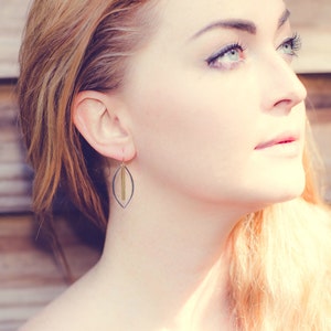 May include: A close-up of a woman wearing a pair of earrings. The earrings have a dark gray, leaf-shaped frame with a vertical gold bar in the center. The earrings are hanging from a gold hook. The background is a blurred wooden surface.