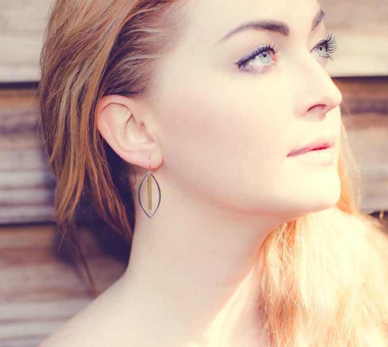 May include: A close-up of a woman wearing a pair of earrings. The earrings have a dark gray, leaf-shaped frame with a vertical gold bar in the center. The earrings are hanging from a gold hook. The background is a blurred wooden surface.