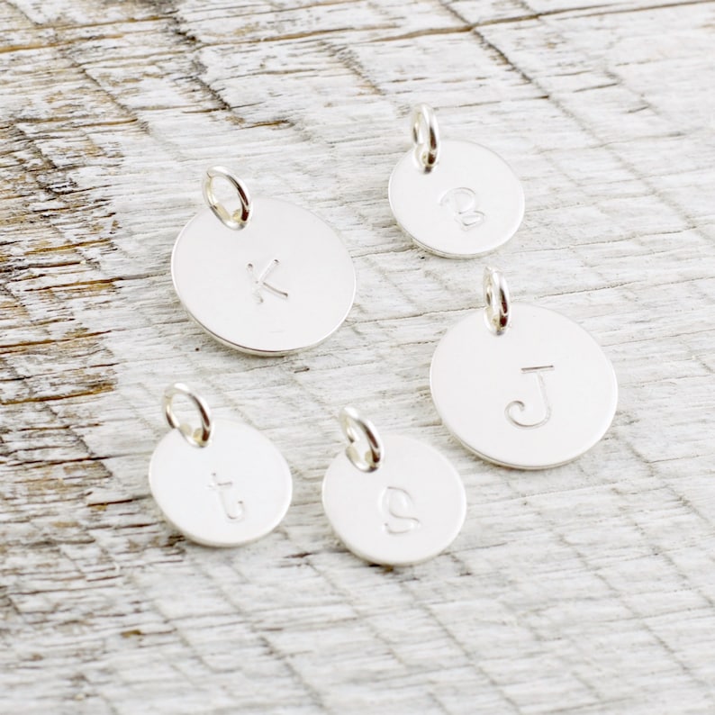 May include: Five silver charms with different letters engraved on them. The letters are K, B, J, t, and g.